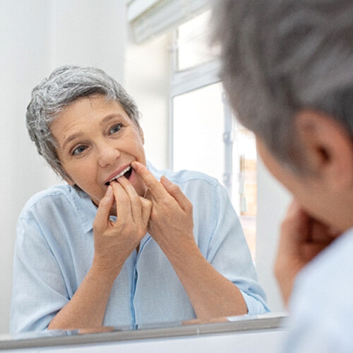 A middle-aged woman flossing around her dental implants while standing in front of the mirror