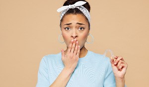 Woman in t-shirt and hair bandana holding Invisalign in one hand covering mouth as if to say “oops” with the other