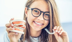 Girl in glasses holding sample braces in one hand and Invisalign in the other