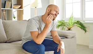 Man with beard rubbing jaw in discomfort while sitting on couch 