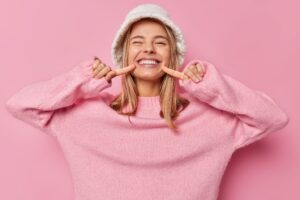 Young woman in pink sweater and white hat pointing to her beaming smile.